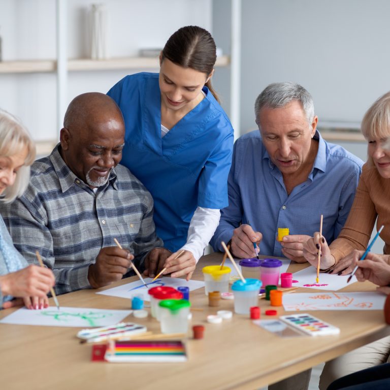 Cheerful creative multiracial senior men and women sitting around table and drawing at nursing home, doing arts and crafts together, young lady nurse assisting group of elderly people, copy space